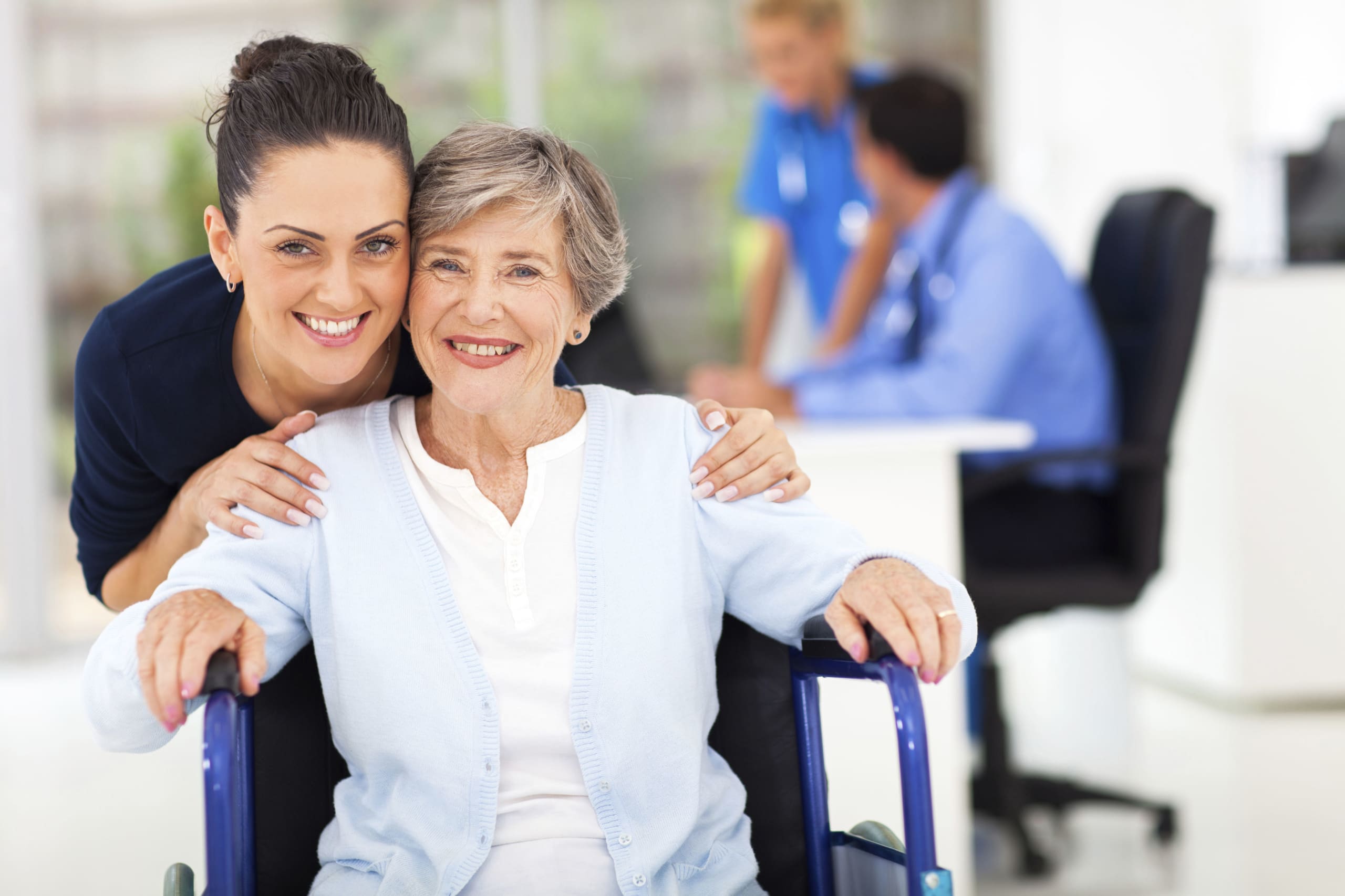 Caregiver smiles beside an elderly woman in a wheelchair, indoors at a care setting.