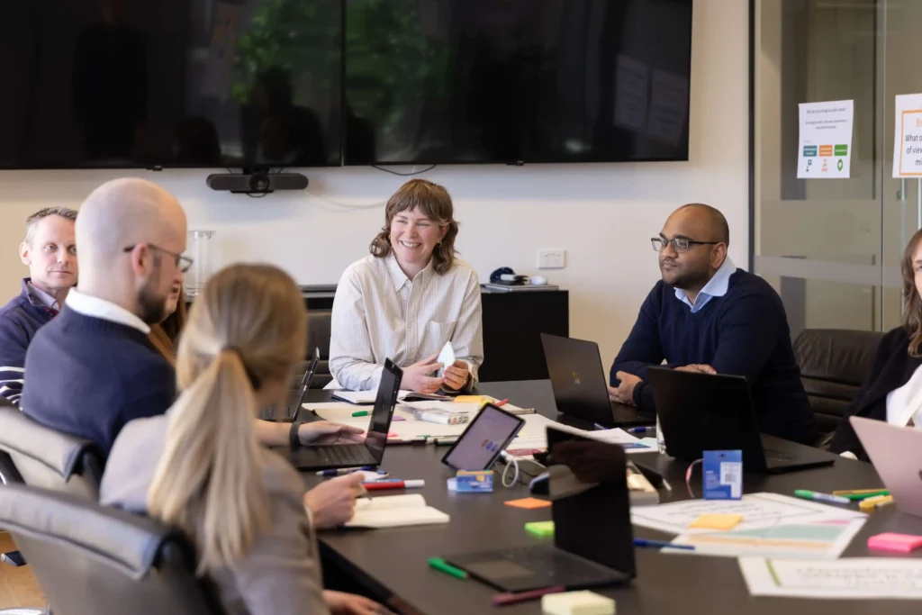 Group of colleagues around a conference table with laptops, notebooks, and sticky notes; the central woman smiles while holding a card/prototype.