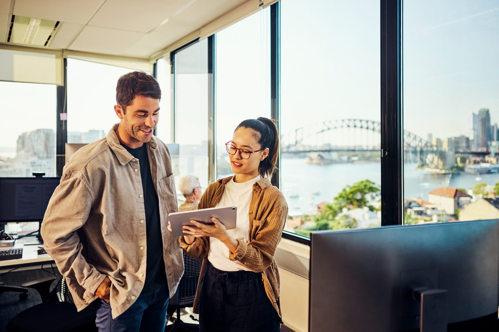 Two colleagues stand by a desk in a bright office, looking at a tablet together with a city and bridge visible through large windows behind them