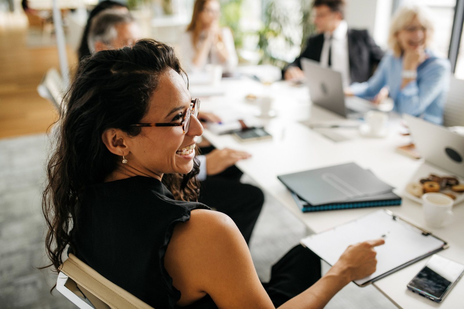 Smiling woman with glasses sits at a conference table, taking notes as colleagues discuss in a bright office. HTML
