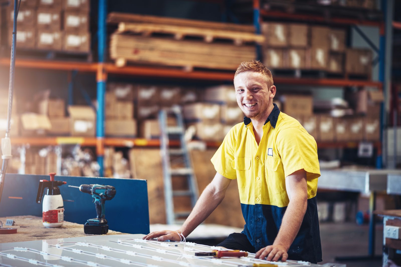 Smiling male worker in a yellow and blue uniform leaning on a worktable in a busy warehouse.