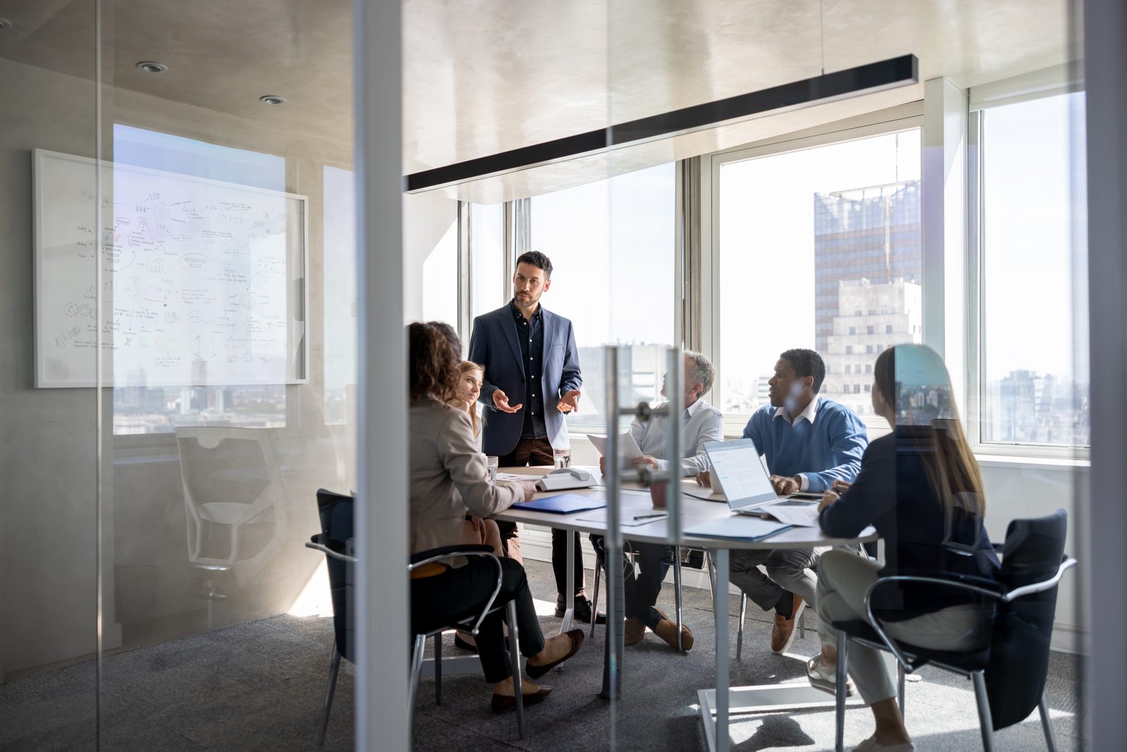 Business meeting in a glass-walled conference room as a man in a blazer presents to colleagues around a table with laptops and papers.