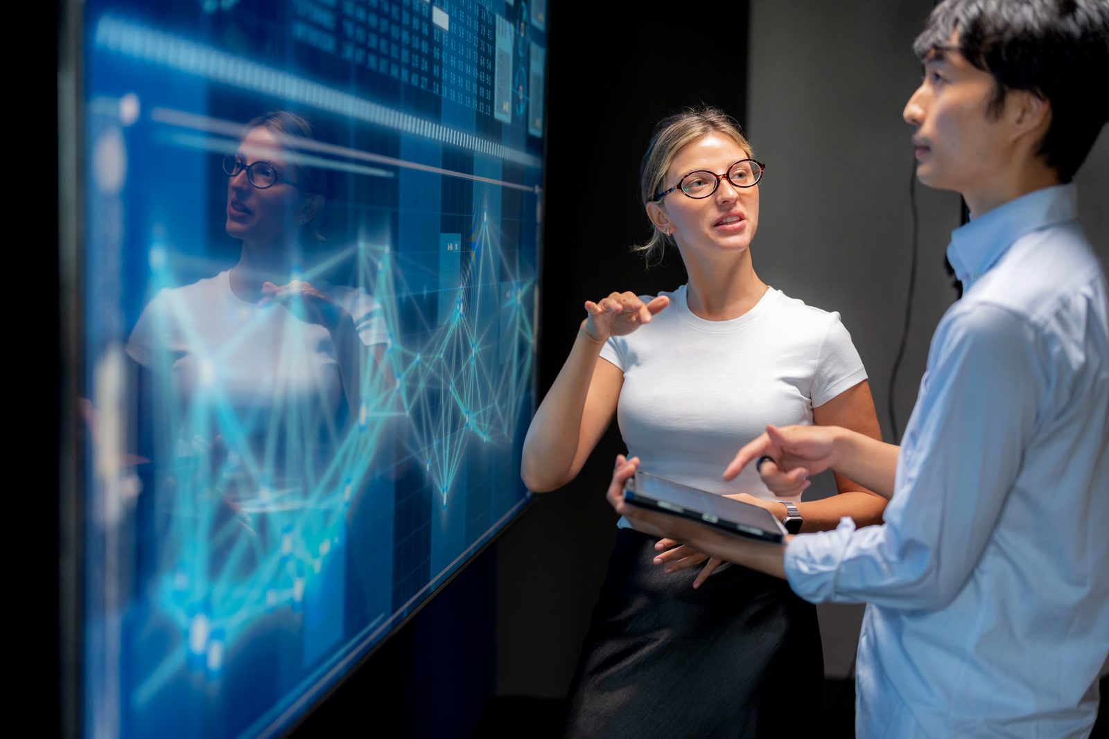 Two professionals discuss data on a large blue touchscreen wall, one gesturing while the other holds a tablet.