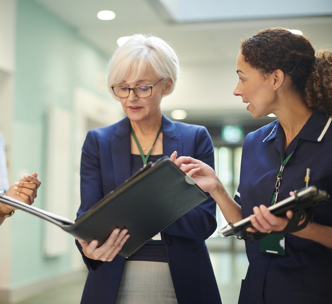 Three healthcare professionals in a hospital corridor discuss a black folder, with stethoscope visible on the left.