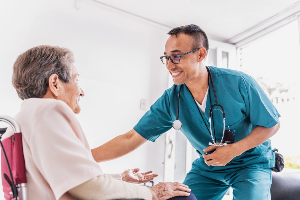 Smiling healthcare worker in teal scrubs greets a seated elderly patient in a wheelchair, offering support and care.