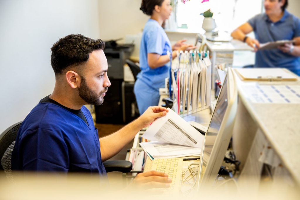 Male medical staff in blue scrubs sits at a computer, reviewing documents at a clinic reception desk with folders nearby