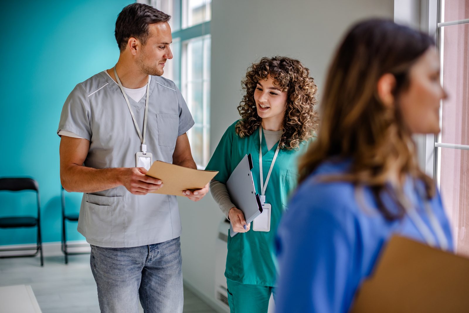 Two healthcare workers in scrubs talk in a bright hospital hallway, reviewing a file folder together after a shift change.