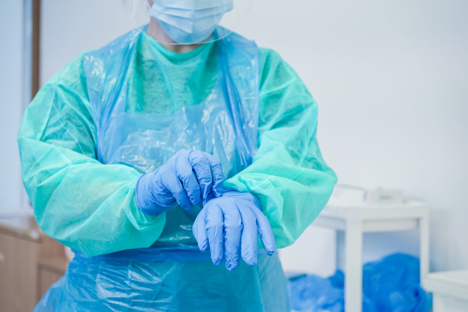 Healthcare worker in full PPE puts on blue gloves, preparing for a procedure