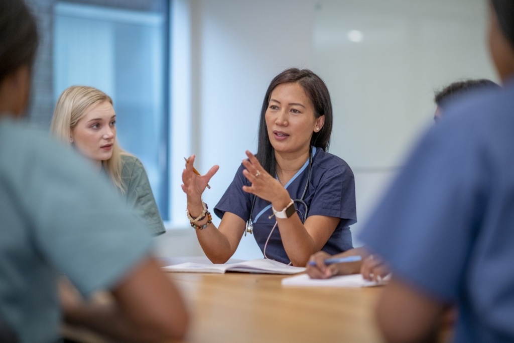 Medical professionals in a conference room discuss patient care as a nurse in scrubs speaks with animated hand movements to the group.
