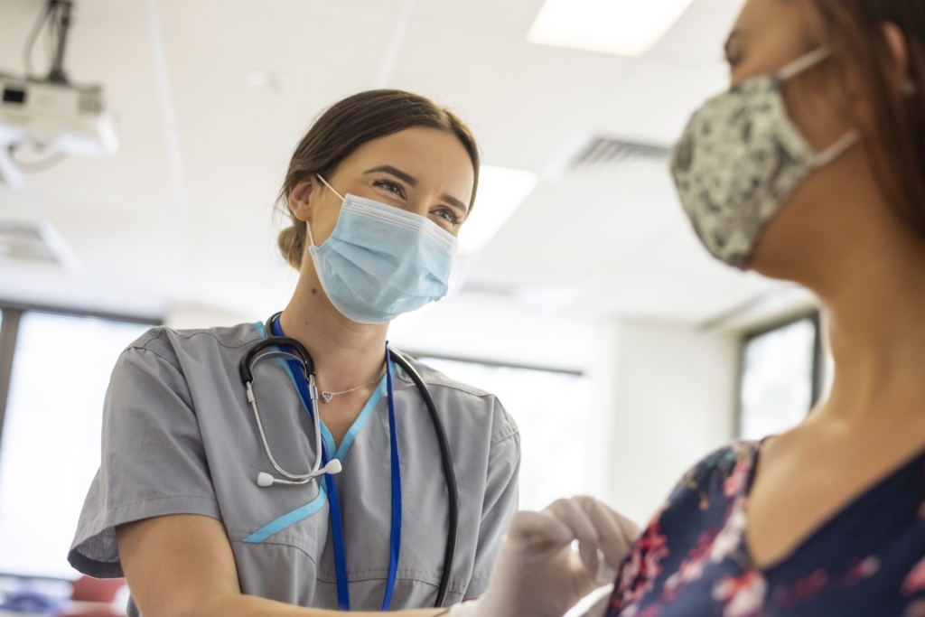 Nurse in scrubs with a stethoscope administers a vaccine to a patient wearing a floral top, both wearing masks in a clinic.