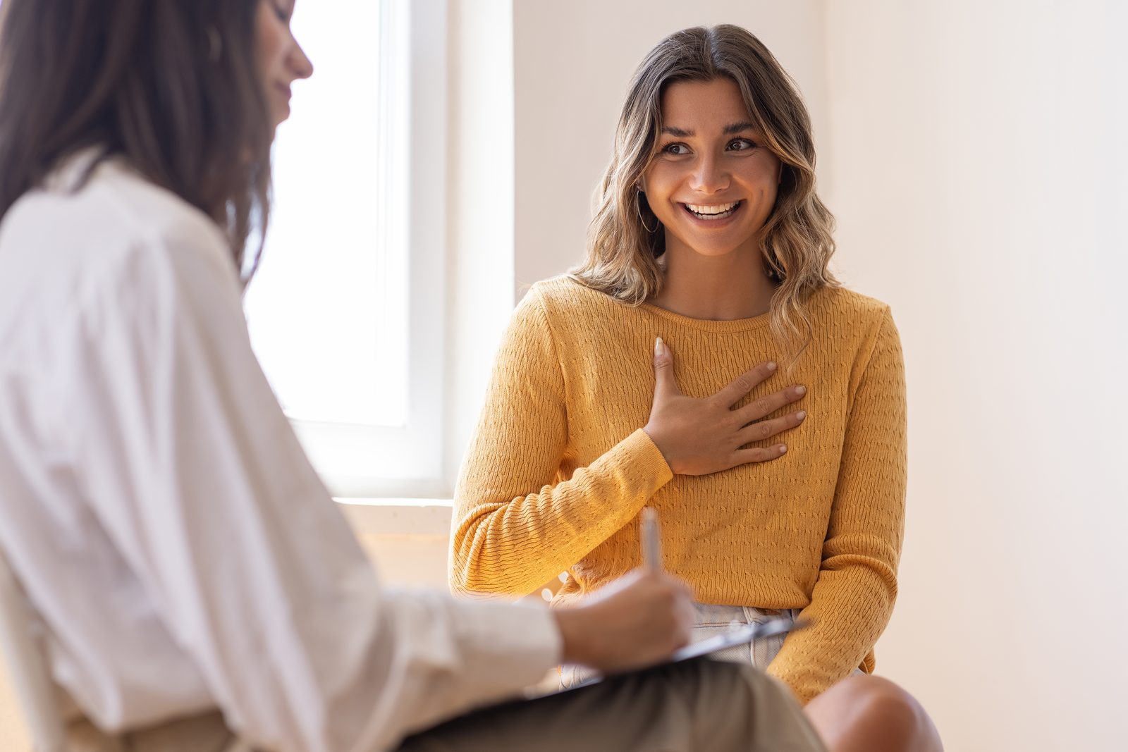 A smiling woman in a yellow sweater sits with her hand over her chest during a counseling session.
