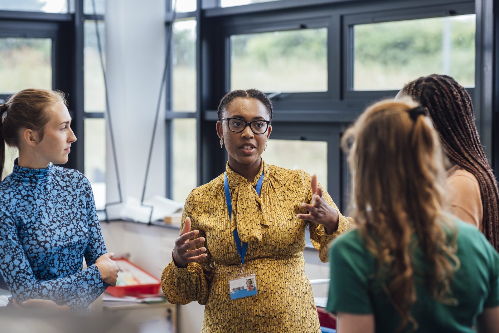 Black woman in a yellow patterned dress and glasses speaks with hand gestures to a group of coworkers in a bright office.