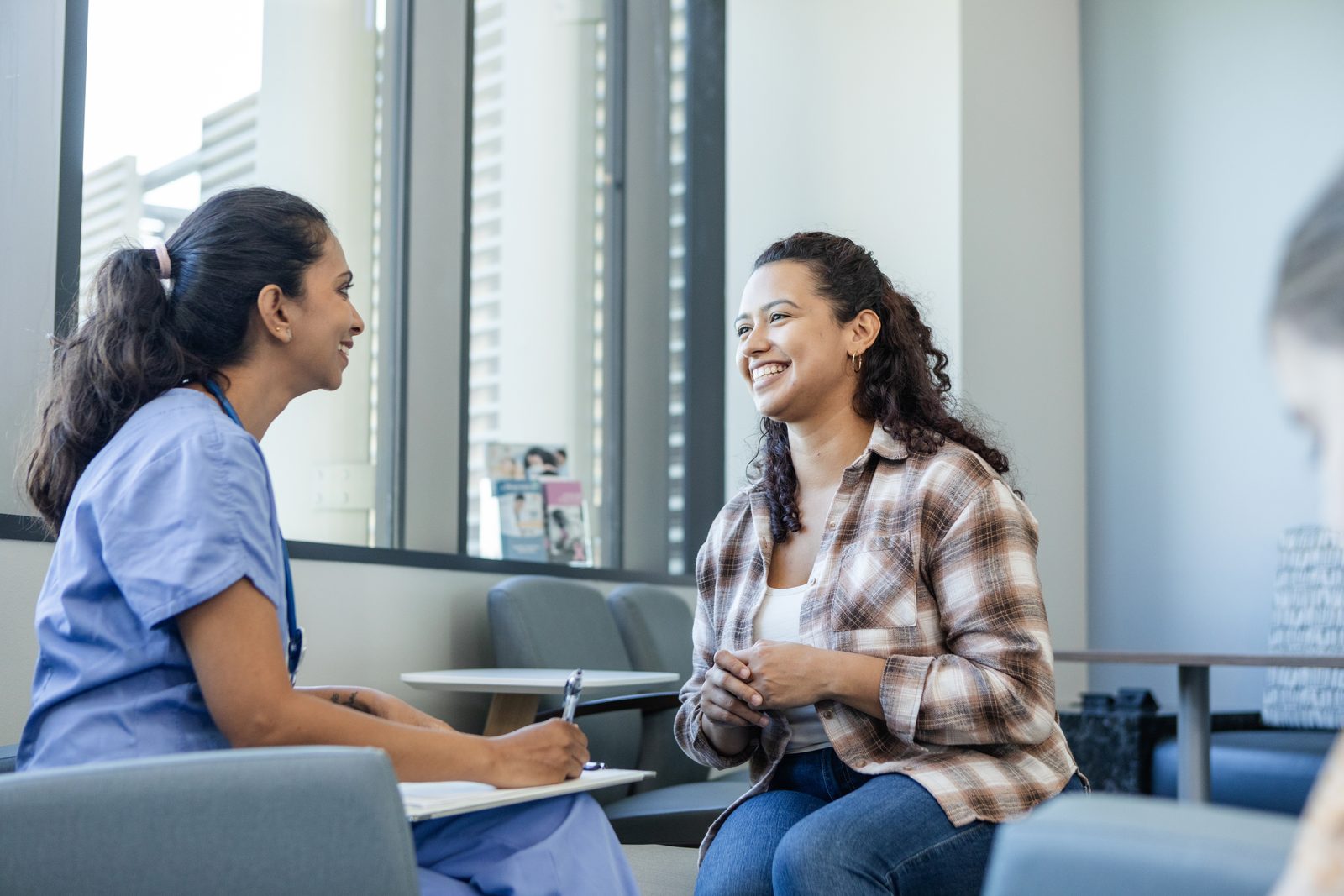 Nurse in blue scrubs chats with a smiling woman in a plaid shirt in a bright waiting area by large windows.