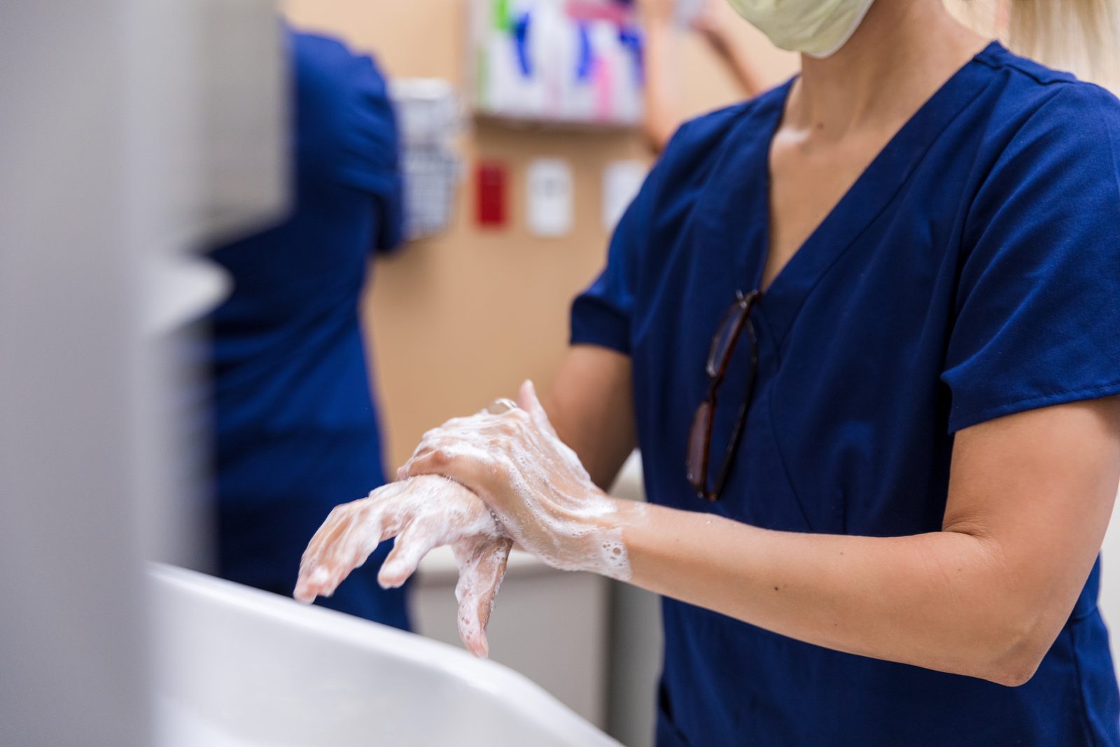 Healthcare worker in blue scrubs and a mask washing hands with soap at a sink, hands covered in lather.