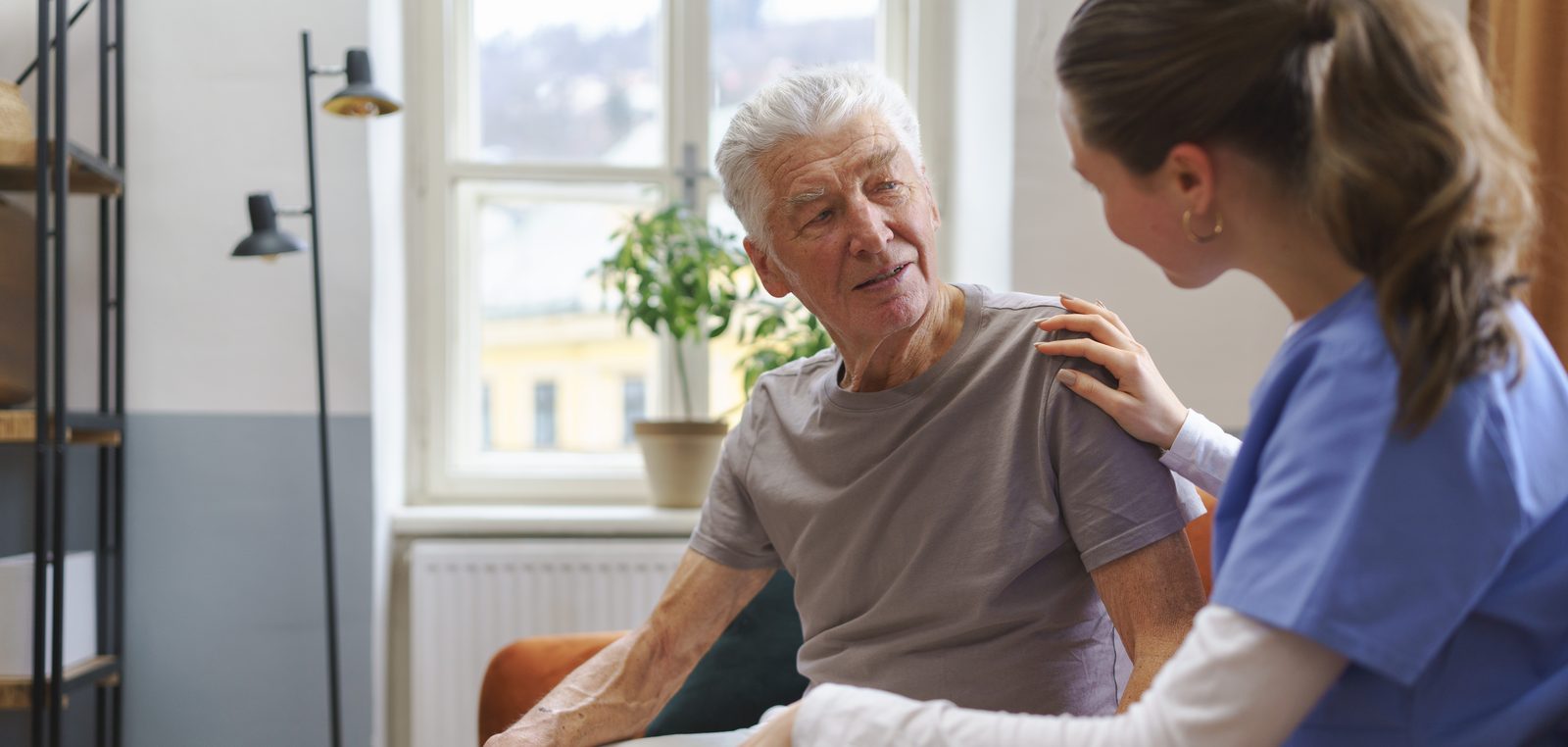Older man talking with a caregiver in a home setting, the caregiver resting a reassuring hand on his shoulder.