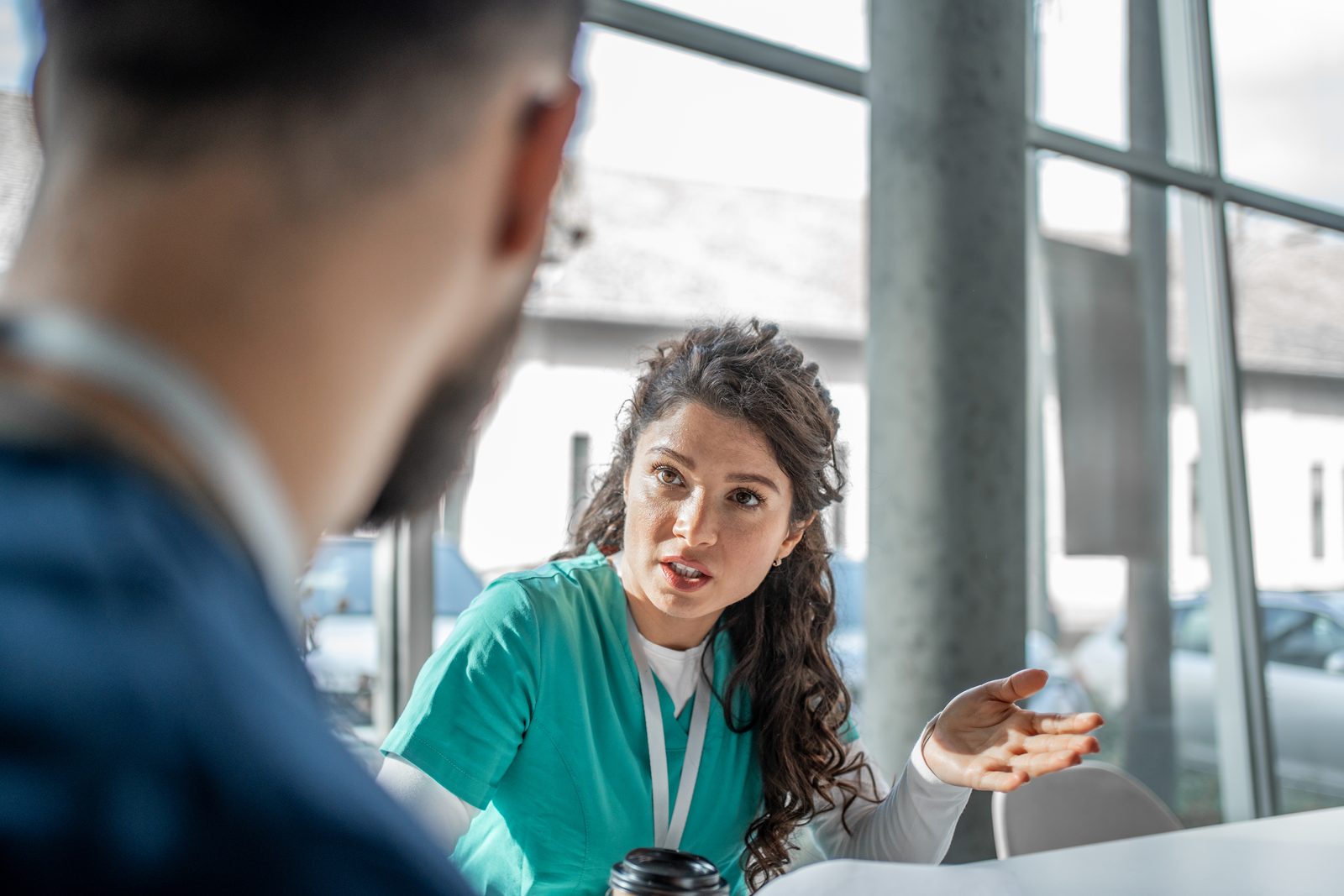Healthcare professional in teal scrubs explains something during a discussion with a patient in a bright, glass-walled setting; her hand is raised as she speaks.