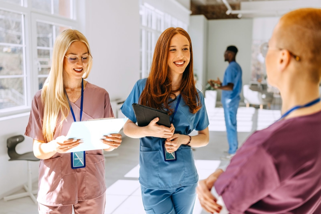 Two female healthcare workers in scrubs smile and chat in a bright hospital hallway, one holding a clipboard and the other a tablet/folio with badges visible.