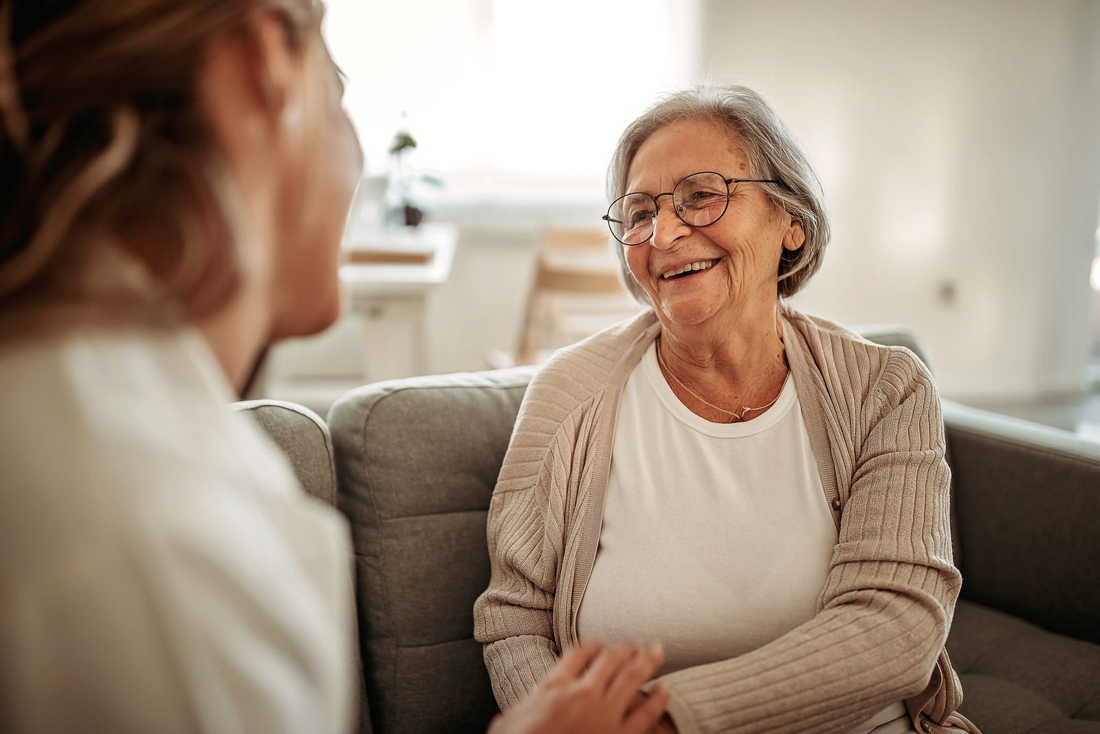 Smiling elderly woman with glasses on a sofa, talking with a caregiver nearby in a bright living room.