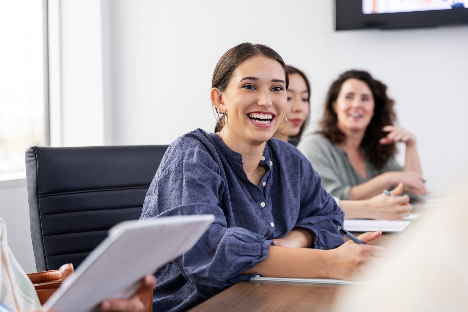 Coworkers at a conference table, with a smiling woman in a blue blouse in the foreground.