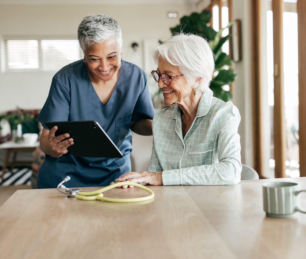 Nurse in blue scrubs shows a tablet to an elderly woman at a kitchen table, discussing health information.
