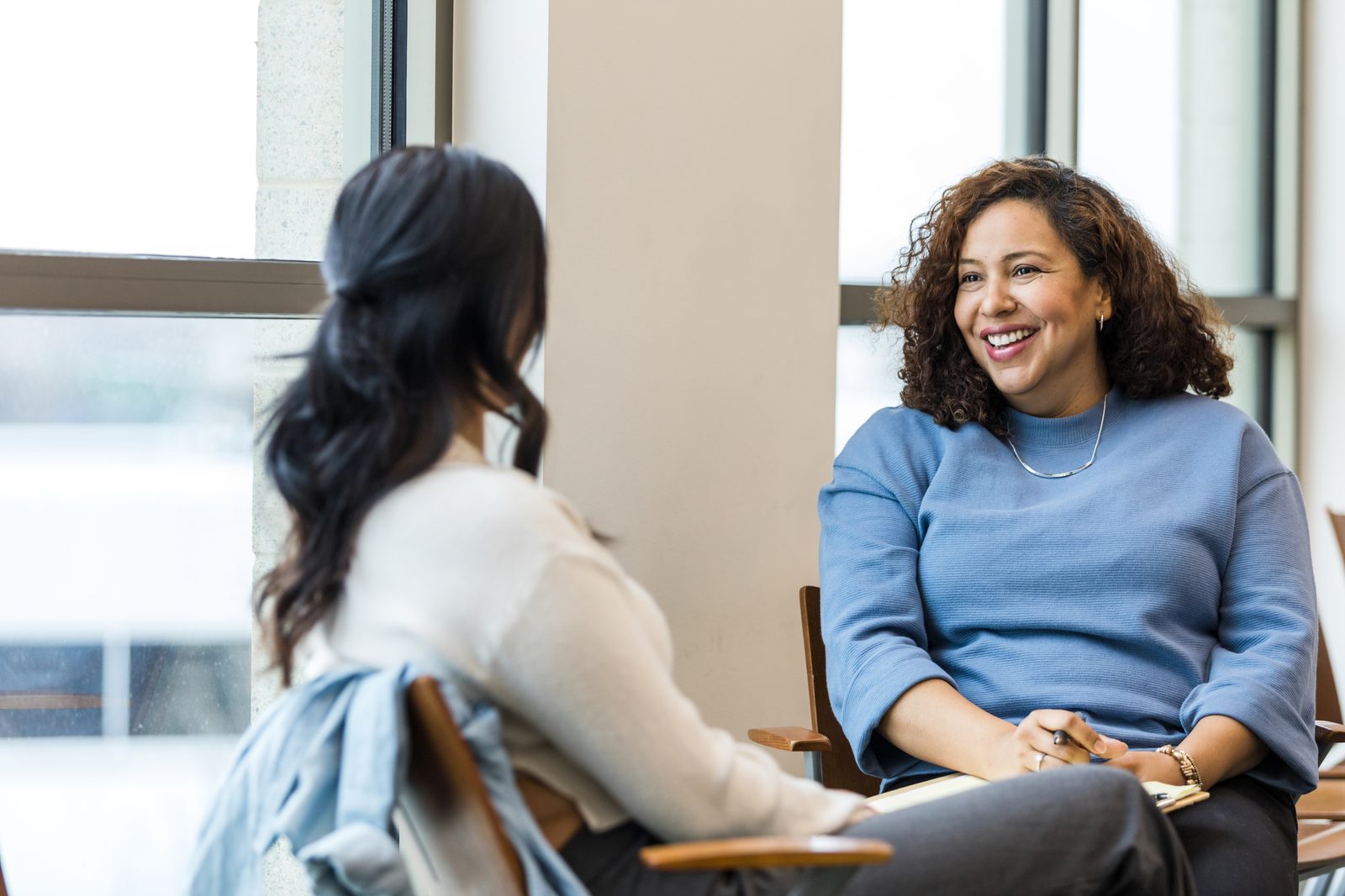 Two women in a bright room chat warmly; one wears a blue sweater and smiles, the other seen from behind in a light top.",