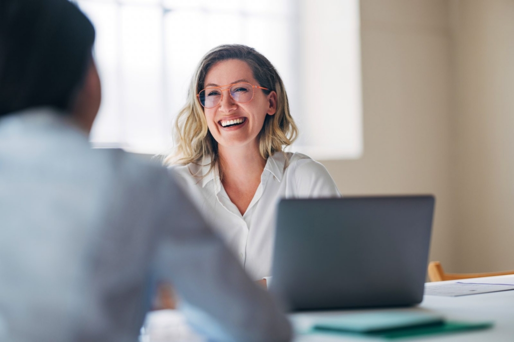 Woman with blonde hair and pink glasses smiles at a coworker across a laptop in a bright office setting.