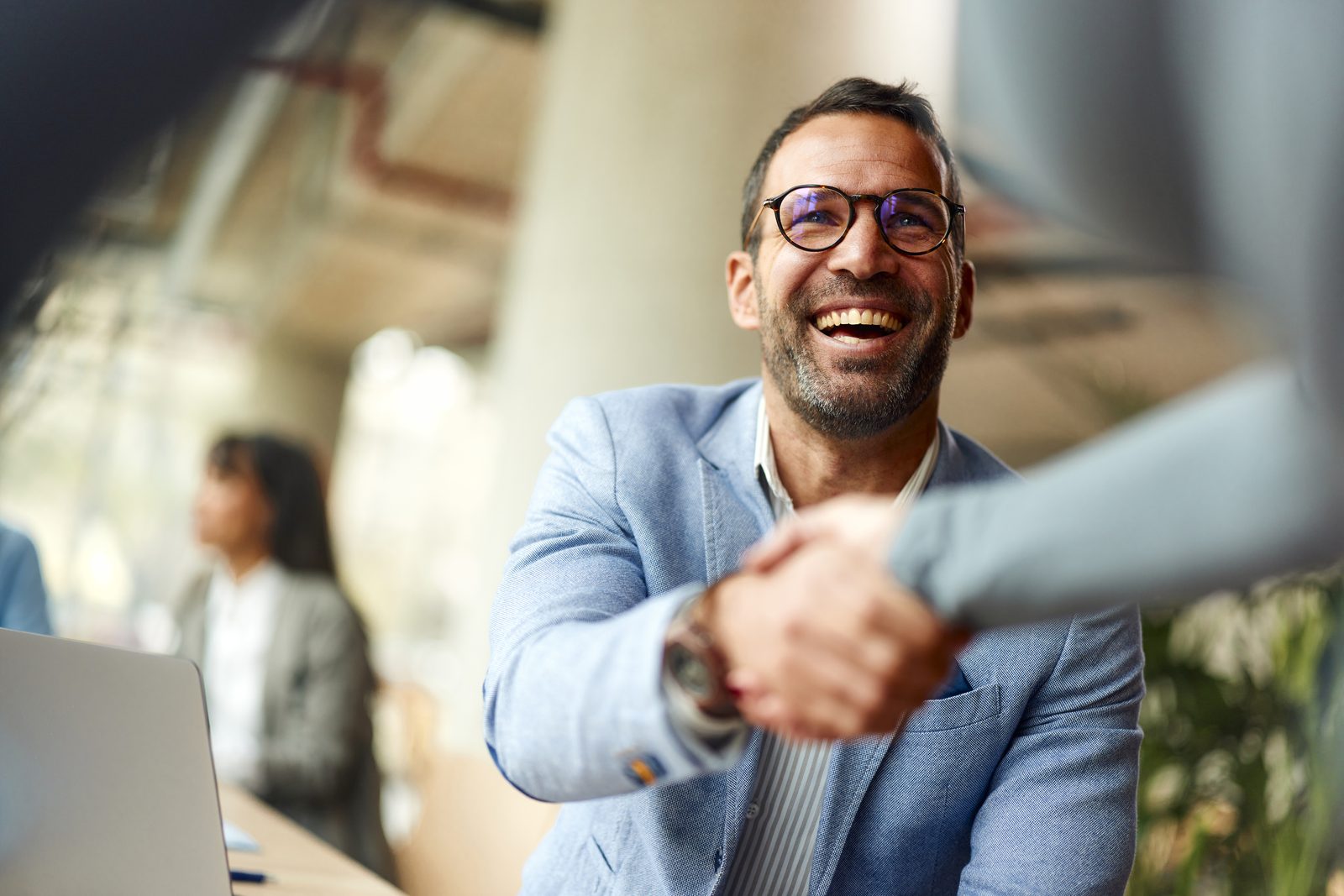Smiling man in a light blazer shakes hands with a coworker in a bright office.