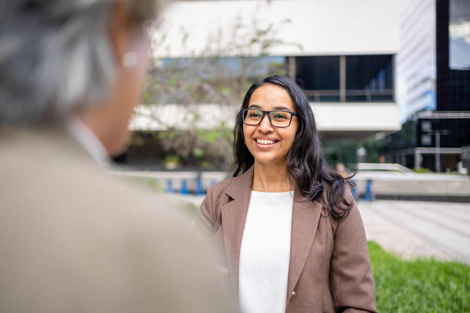 Smiling woman with glasses and a brown blazer talks with a man outdoors on a campus courtyard.