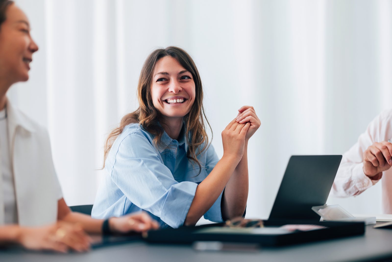 Smiling woman in a blue shirt sits at a conference table with a laptop, hands clasped in front.