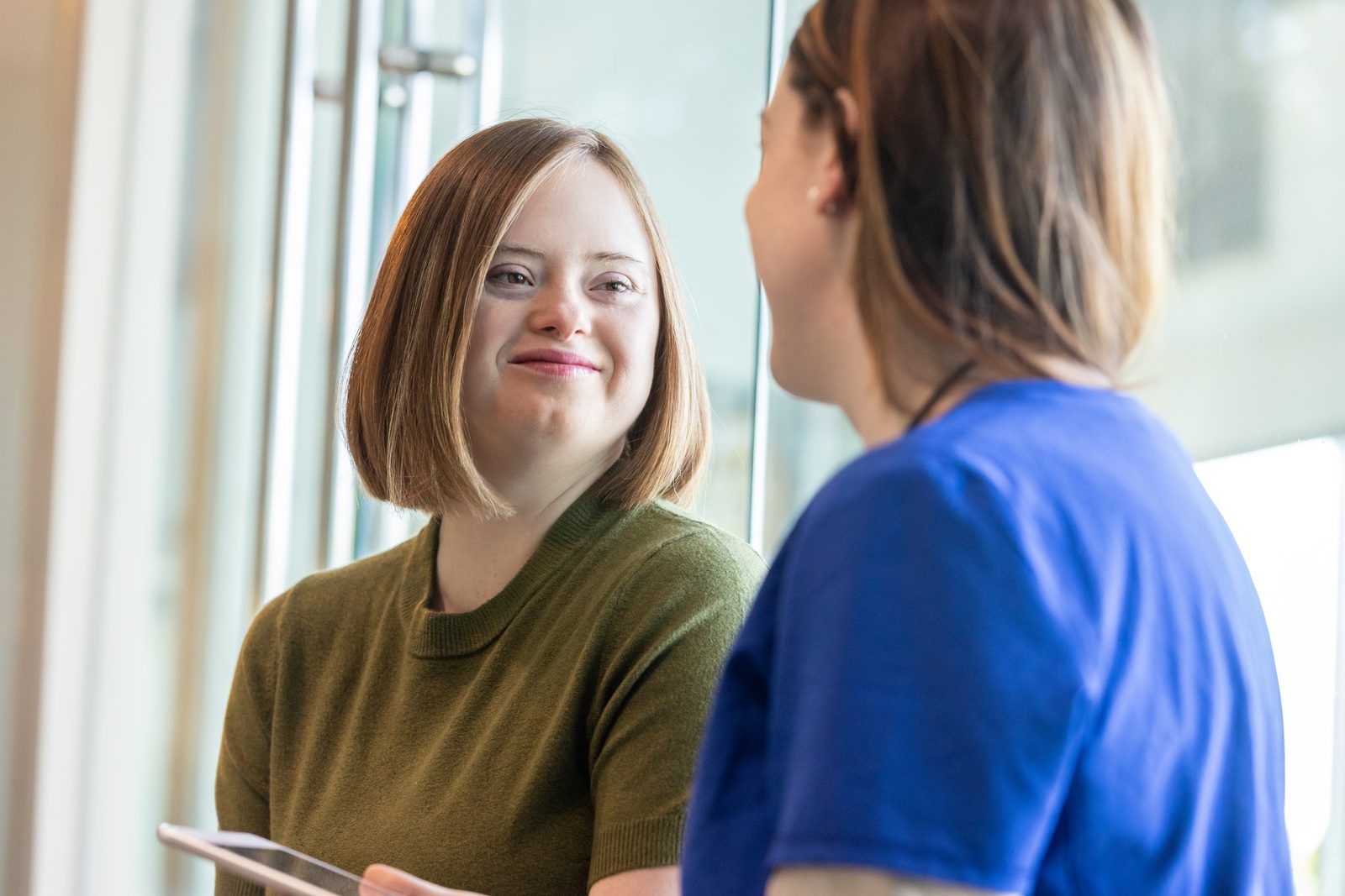 Smiling woman in a green sweater holds a tablet and chats with a colleague in blue scrubs near a bright window.