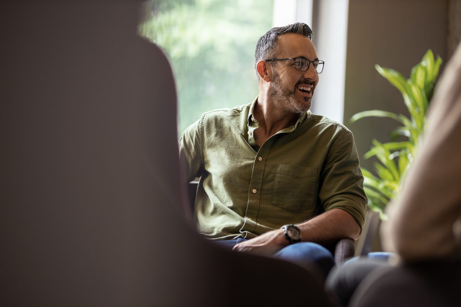 Man with glasses and a gray beard, wearing a green shirt, smiling while talking in a casual office meeting.