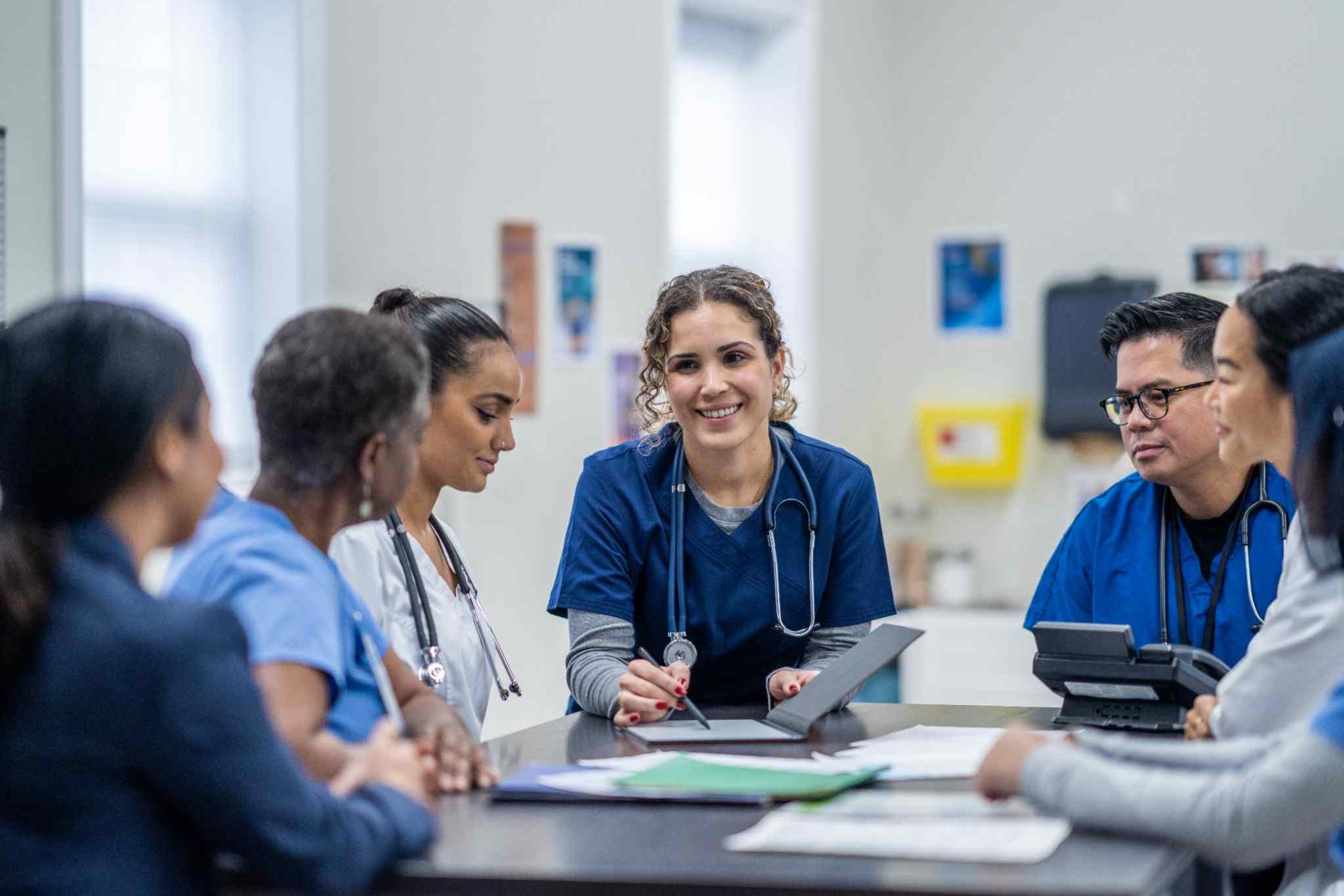 Medical staff in blue scrubs and white coats gather around a table, reviewing documents together in a clinical setting.