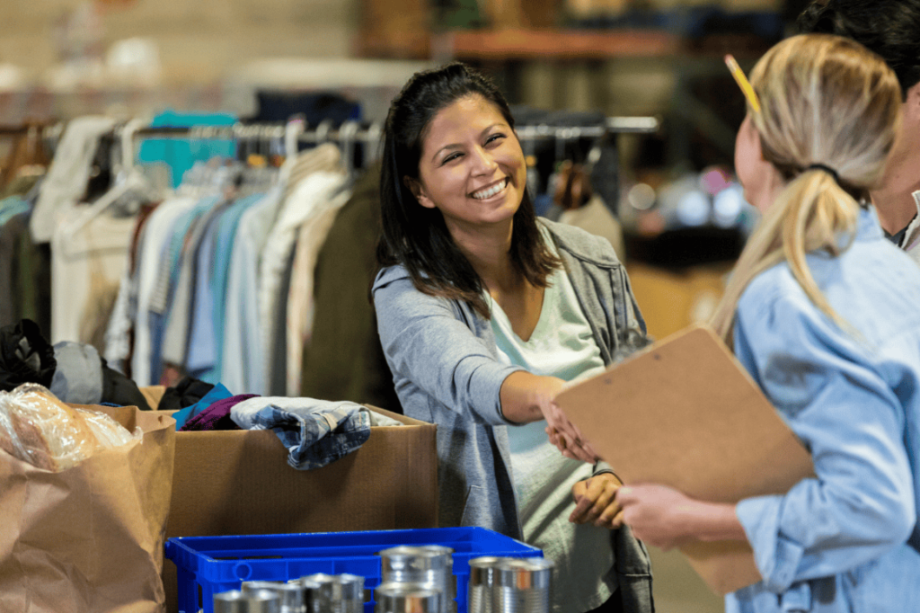 Smiling woman in a gray cardigan hands a clipboard to a customer in a thrift store.