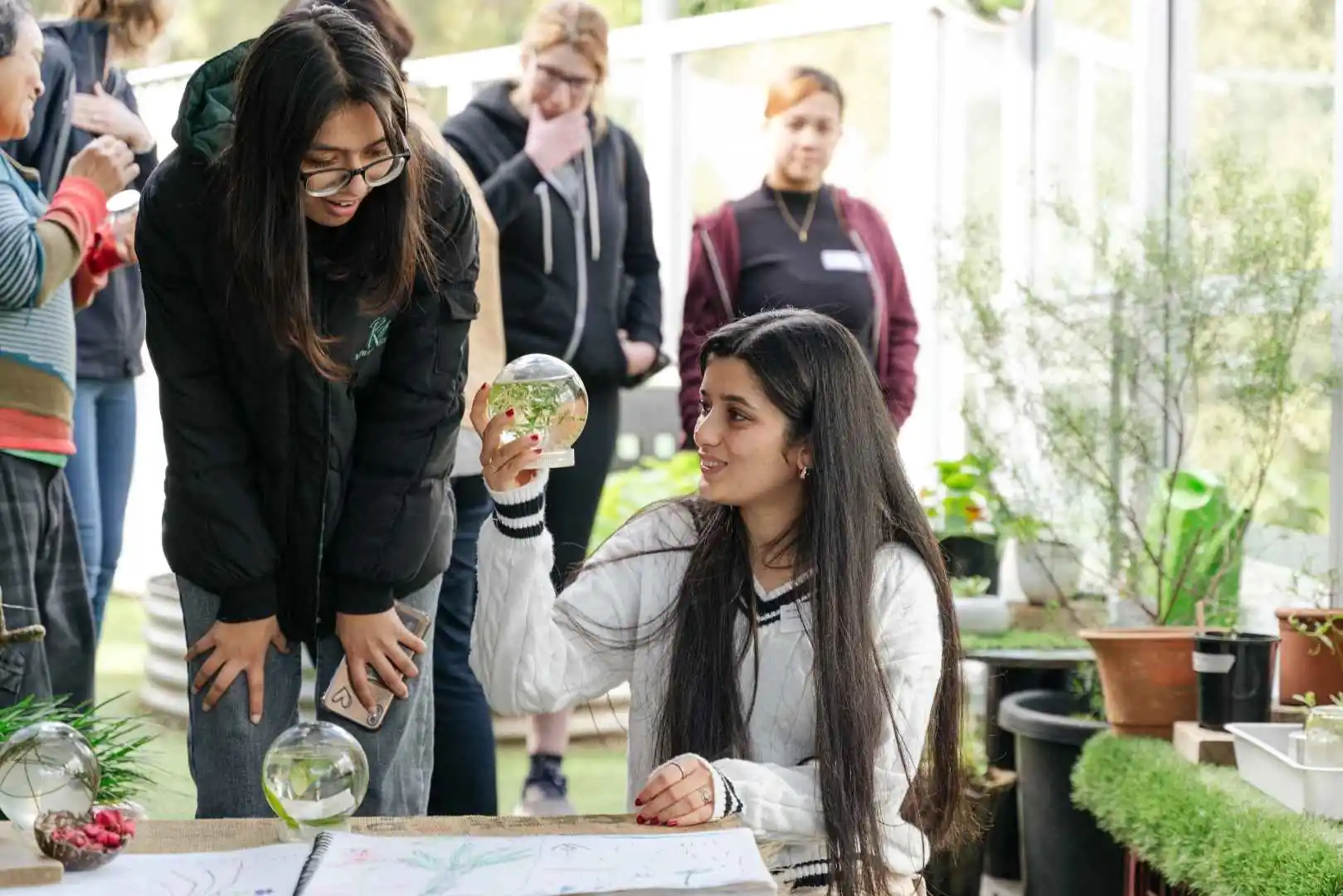 Woman kneeling, holding a glass terrarium globe, smiling at a seated friend in a bright plant-filled room.