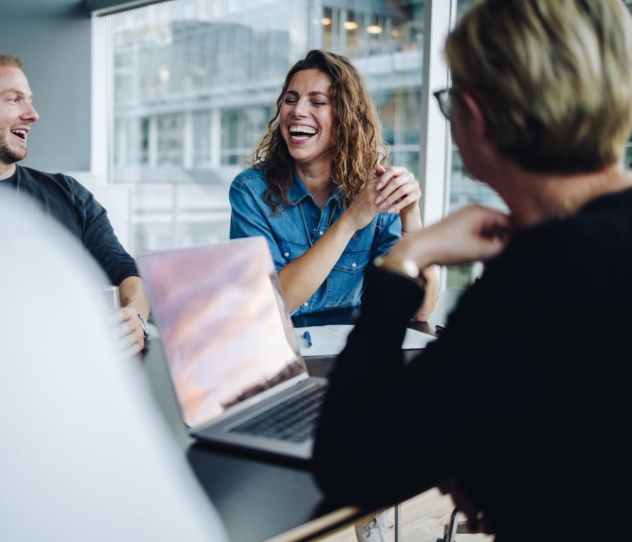 Three colleagues in a bright office laughed during a work meeting around a laptop on the table.