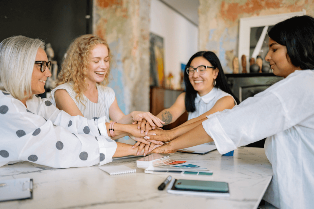 Group of four women of diverse ages join hands on a table, smiling in a collaborative team moment.