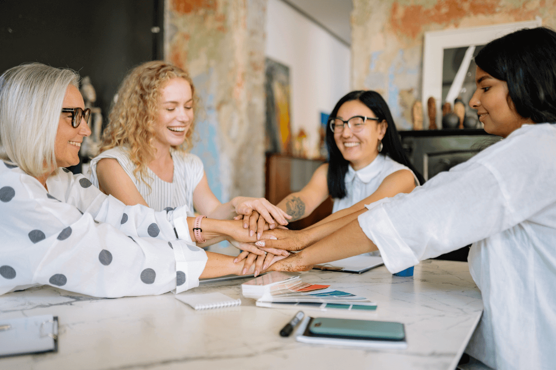 Group of four women of diverse ages join hands on a table, smiling in a collaborative team moment.