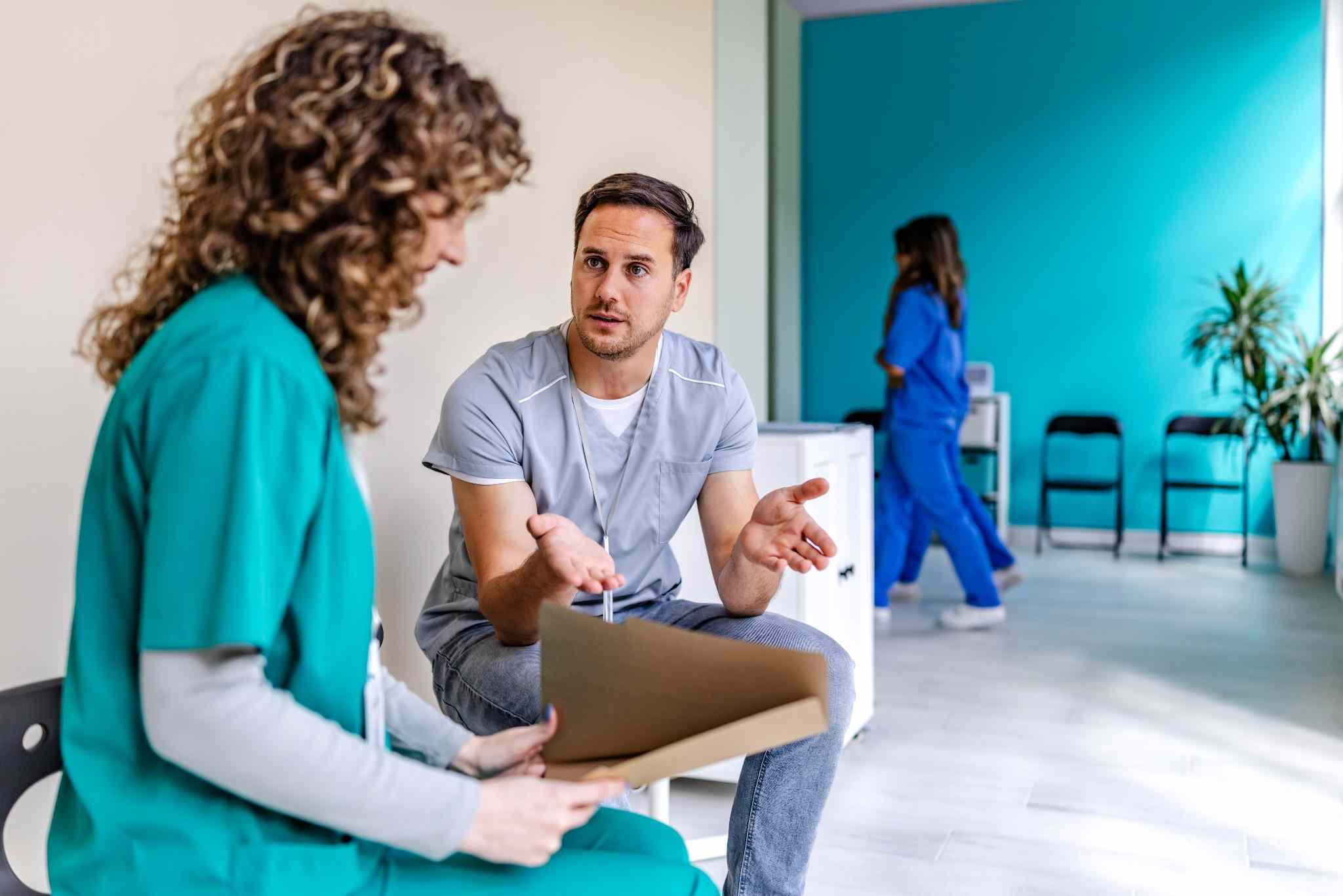 Healthcare worker in gray scrubs talks with a nurse in teal scrubs, holding a folder in a bright clinic lobby with a teal wall in the background.