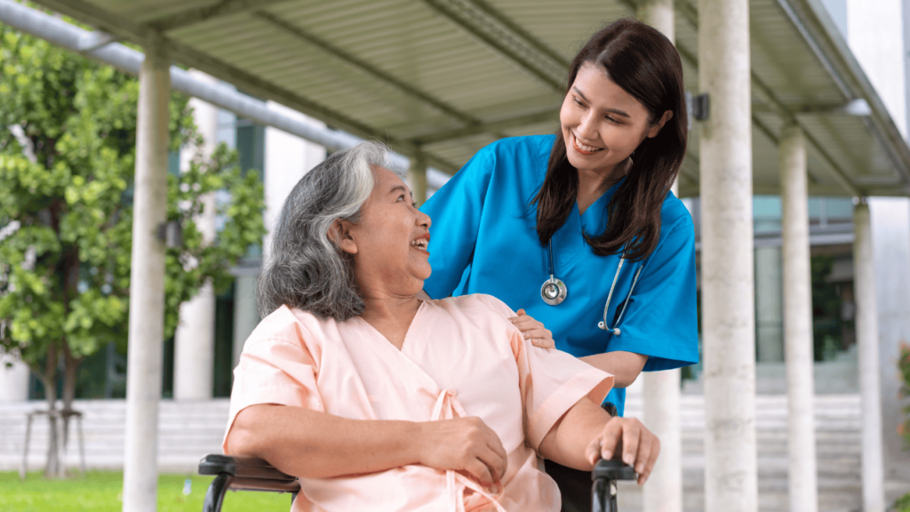 Nurse in blue scrubs smiles at an elderly woman in a wheelchair outdoors.