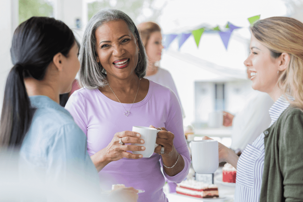 Women chatting and smiling at a casual get-together, holding white mugs, with cake on the table behind them.