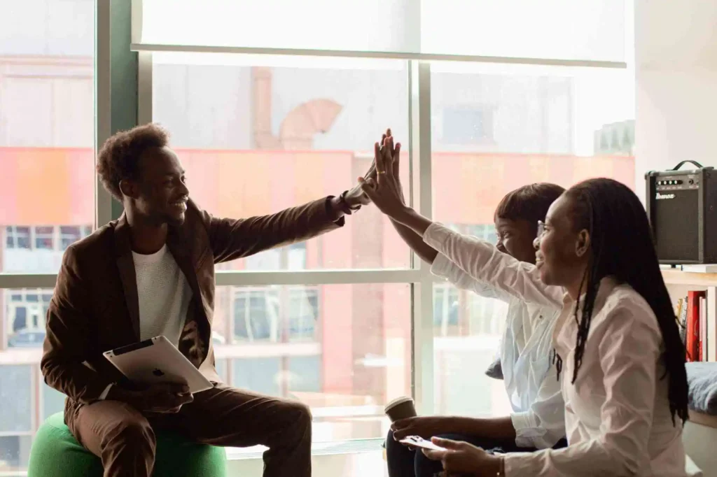 Three coworkers celebrate with a high-five in a bright office; a man holds a tablet while a woman sits nearby and another woman joins the high-five by the window.