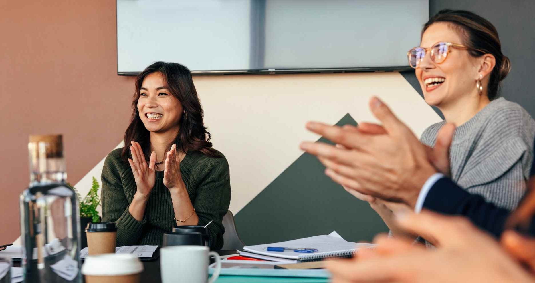 Group of colleagues at a meeting clapping and smiling, with notebooks and coffee cups on the table in a bright office.