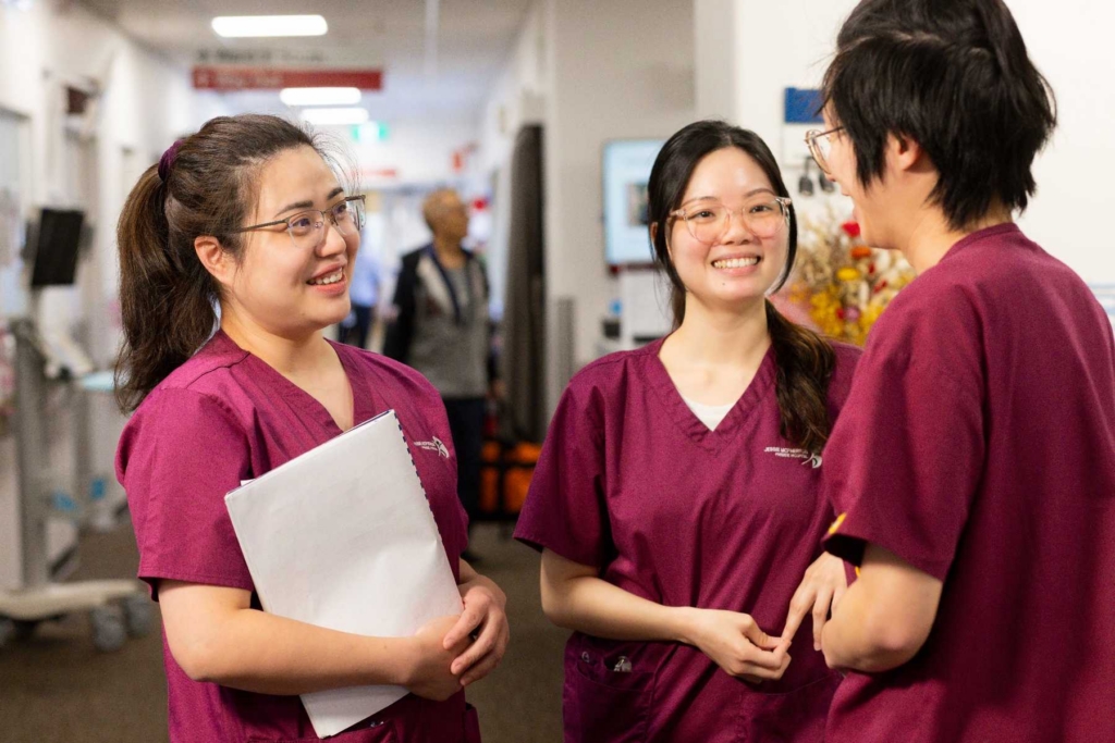 Three healthcare workers in maroon scrubs chat and smile in a hospital hallway; one woman holds a clipboard.