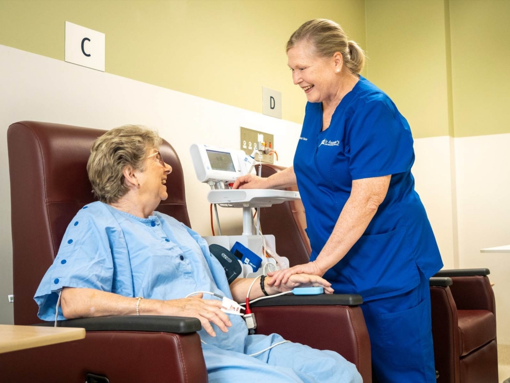 Nurse in blue scrubs takes a patient’s blood pressure while the patient sits in a medical chair and smiles.