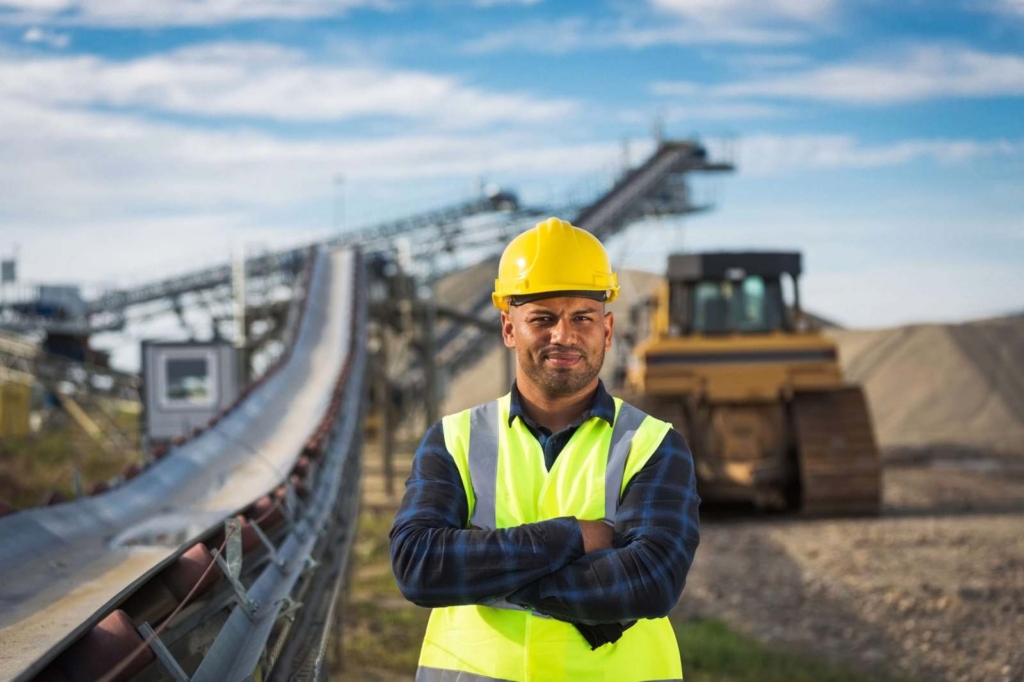 Construction worker in a high-visibility vest and yellow hard hat stands with arms crossed at an industrial site under a conveyor belt and excavator in the background