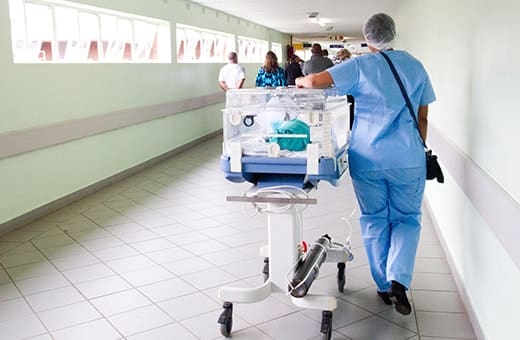 Nurse in blue scrubs pushes a newborn incubator on a wheeled cart down a hospital hallway as staff walk in the background.