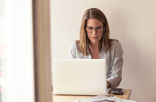 Woman with glasses sits at a desk typing on a white laptop, with papers and a smartphone nearby.