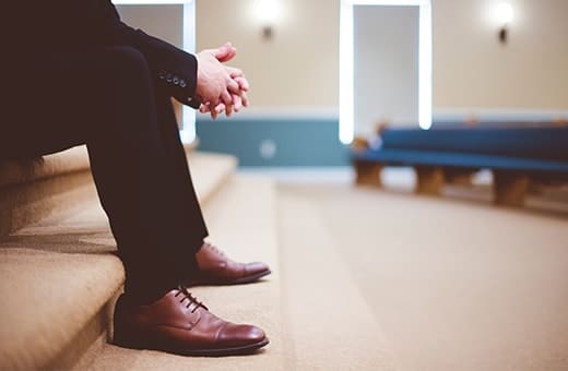Man in a dark suit sitting on church steps, hands clasped, brown dress shoes visible.