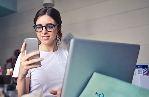 Young woman with glasses holds a smartphone while working on a laptop at a desk in a bright workspace.