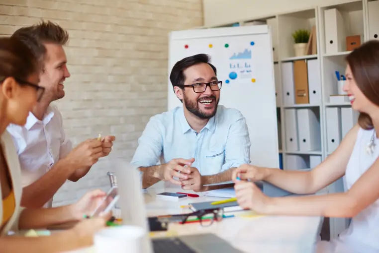 Team of colleagues in a bright meeting room, smiling and collaborating around a table with a whiteboard in the background.